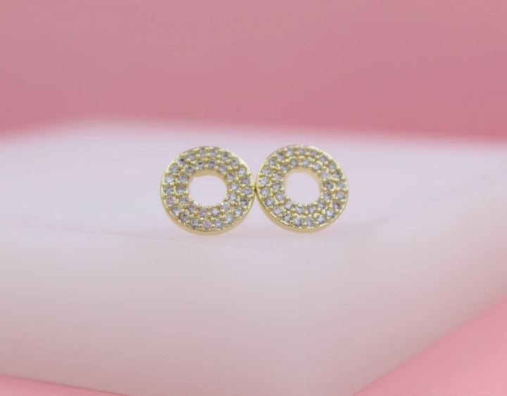Two sparkling gold pave circle stud earrings rest on a white surface against a soft pink backdrop.