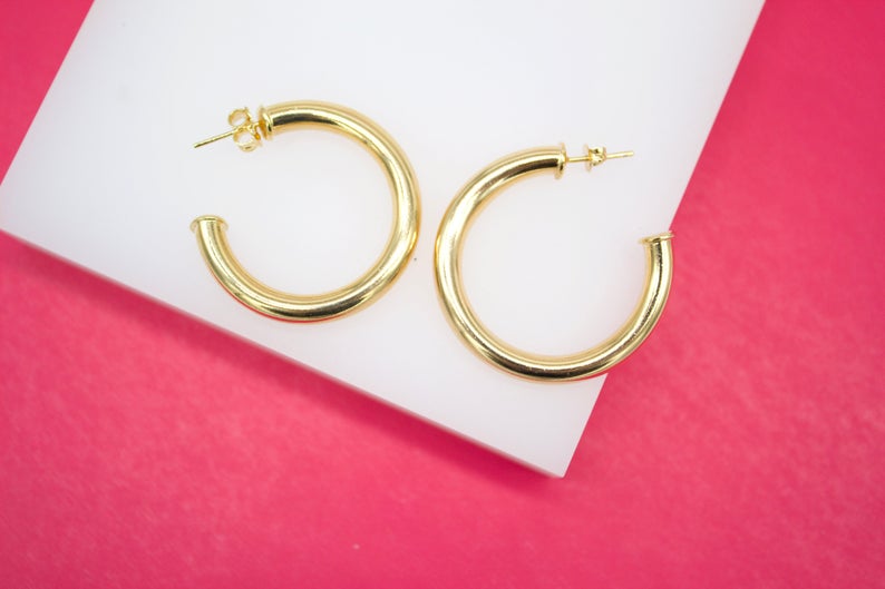 A pair of thick gold hoop earrings rests on a white display block against a vibrant pink background.