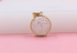 A round gold pendant filled with sparkling loose crystals rests against a pink background.
