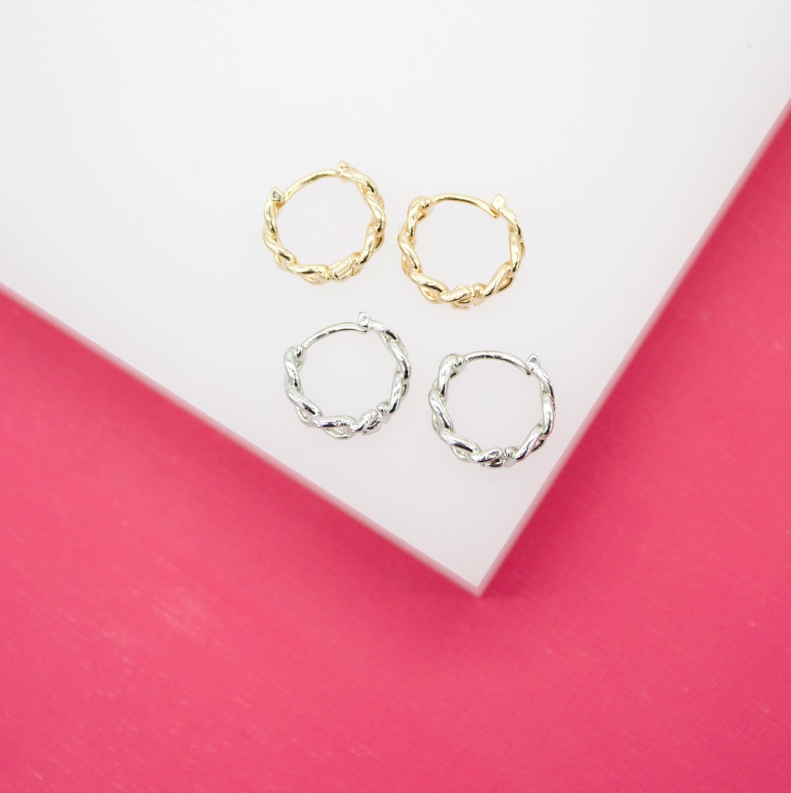 Two pairs of gold and silver twisted hoop earrings rest on a white display block against a vibrant pink background.
