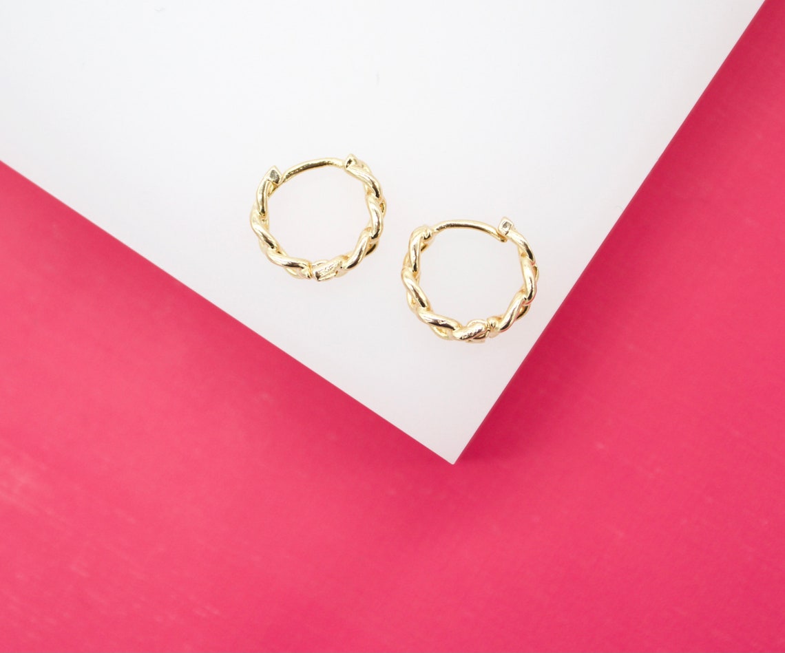 Two gold twist hoop earrings rest on a white surface against a bright pink background.