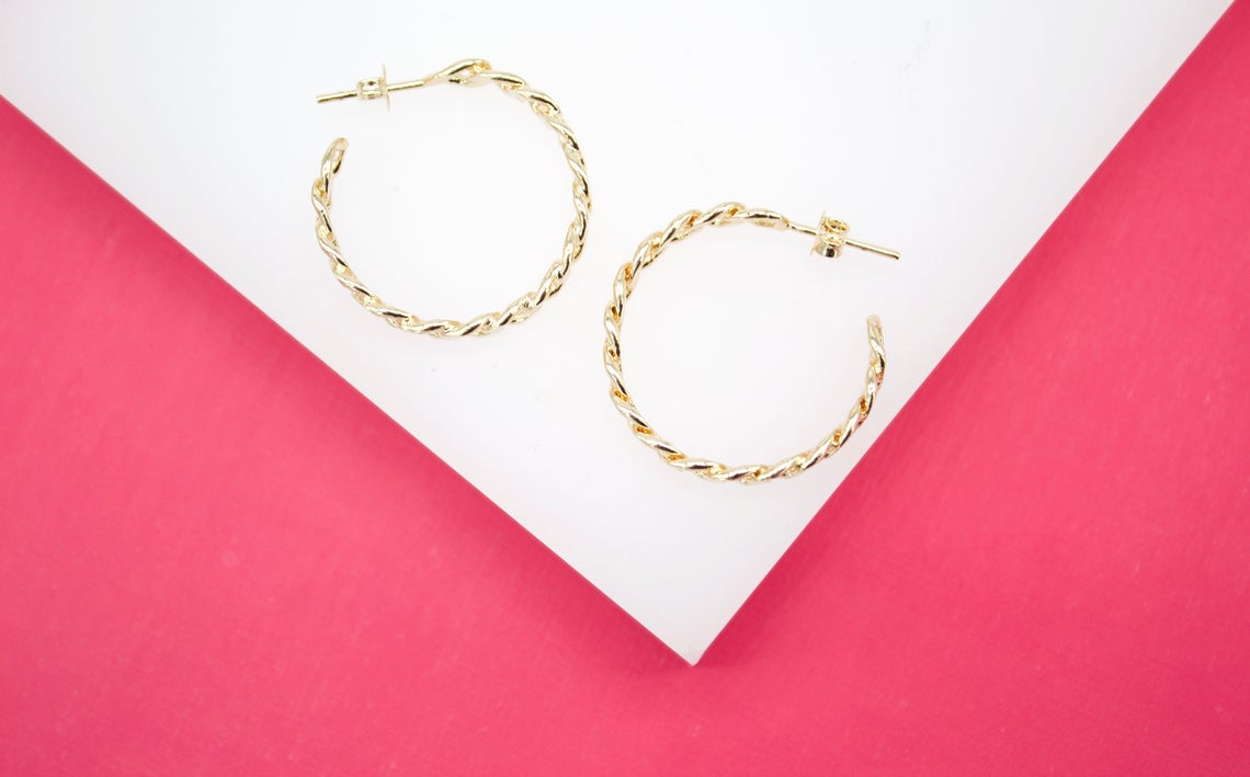 A pair of polished gold twisted hoop earrings rests on a white platform against a vibrant pink background.