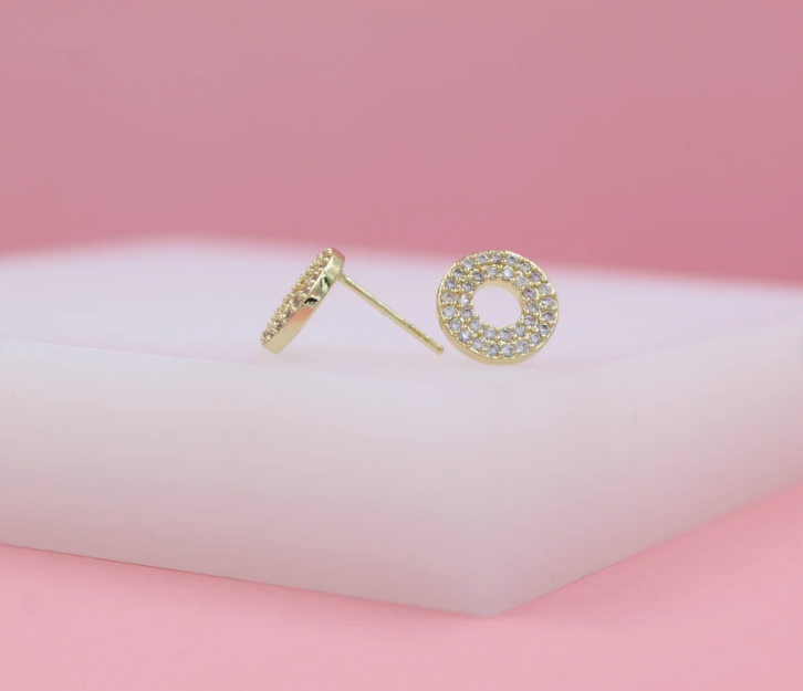 A pair of gold circular stud earrings, encrusted with sparkling cubic zirconia pavé stones, rests delicately on a white display block against a soft pink backdrop.