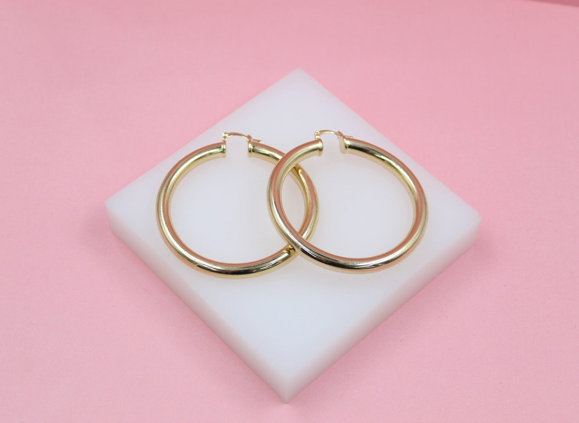 A pair of polished gold hoop earrings rests on a white display block against a soft pink background.