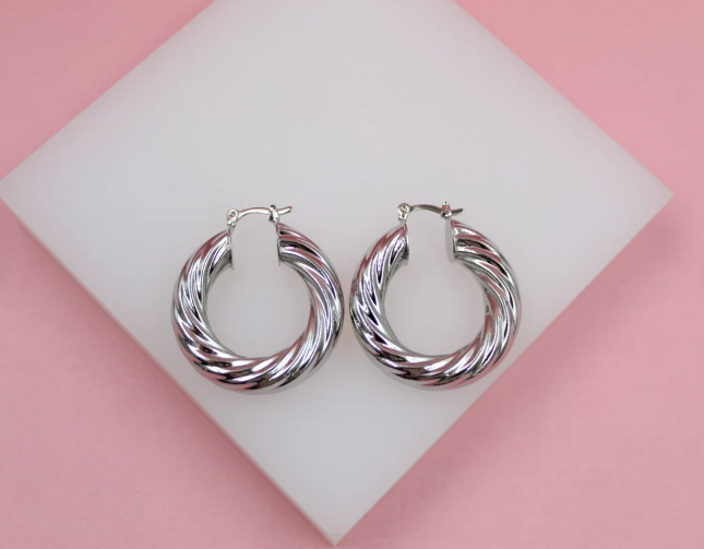 Two shiny silver twist hoop earrings rest on a white display stand against a soft pink background.