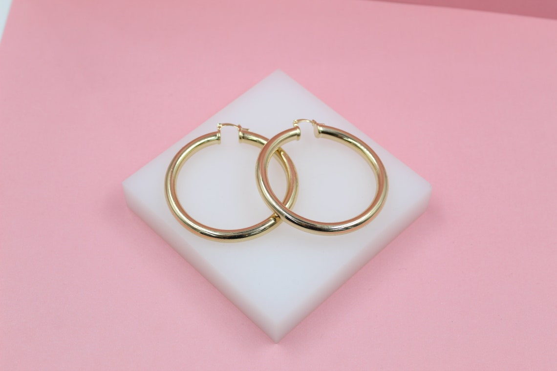A pair of polished gold hoop earrings rests on a small white display block against a soft pink background.