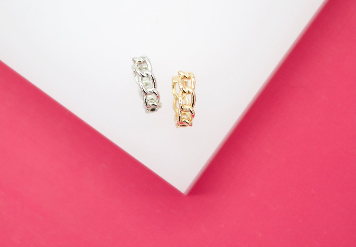 A silver chain link earring and a gold chain link earring rest on a white surface against a pink background.