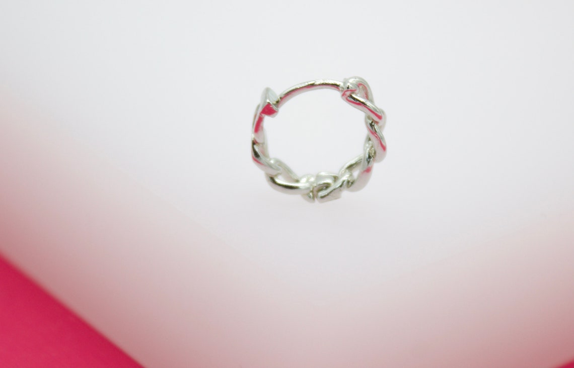 A polished silver huggie hoop earring featuring an intricate twist design rests against a soft white and pink background.