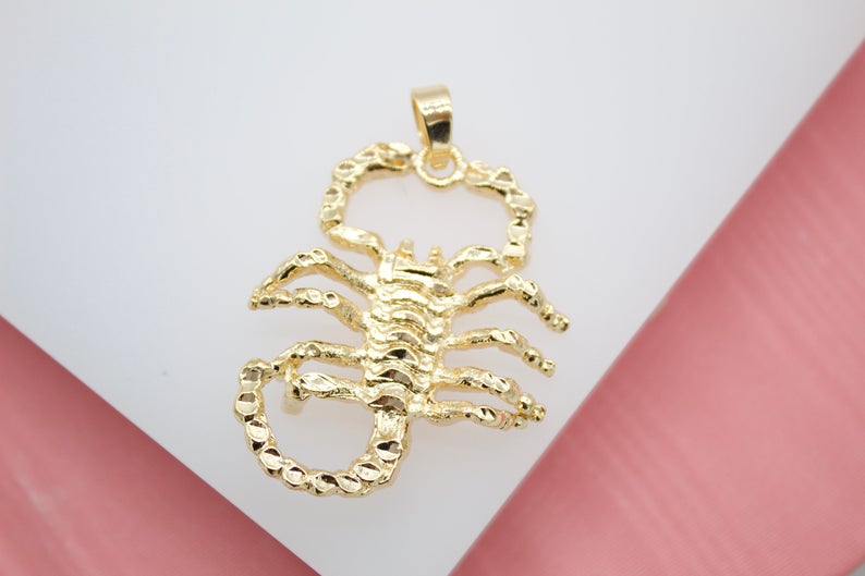 A textured gold scorpion pendant rests on a clean white surface.