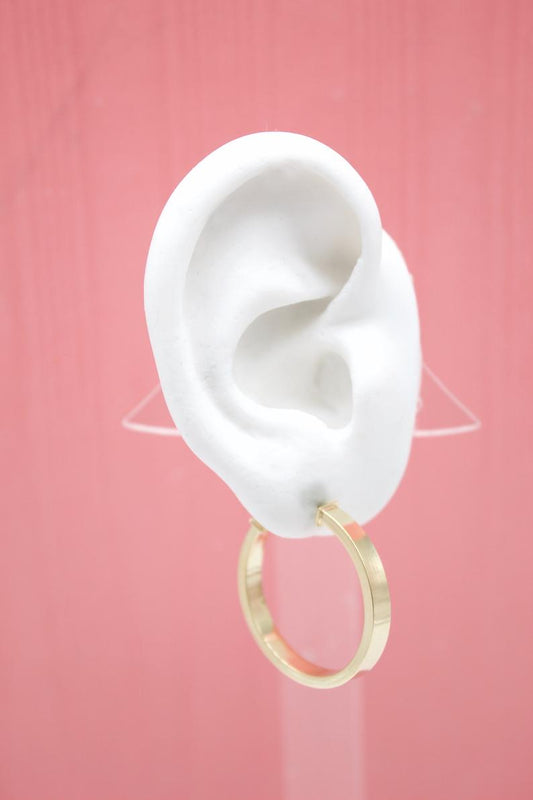 A white mannequin ear displays a modern gold flat-tube hoop earring against a pink background.