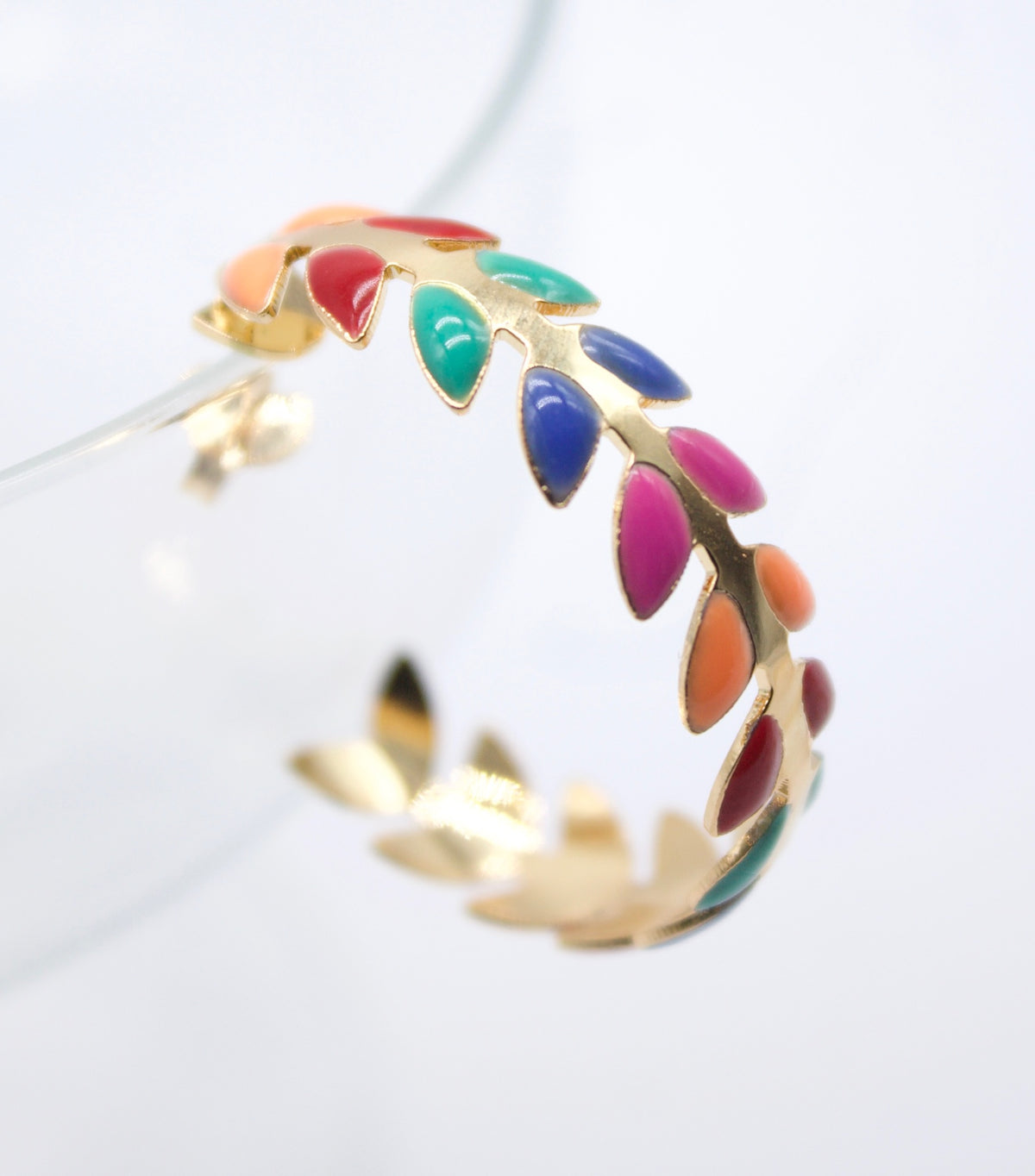 A gold hoop earring features a colorful enamel leaf pattern against a bright white background.