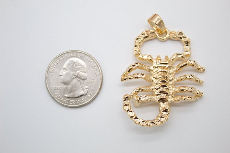 A detailed gold scorpion pendant rests next to a US quarter coin for scale on a white surface.