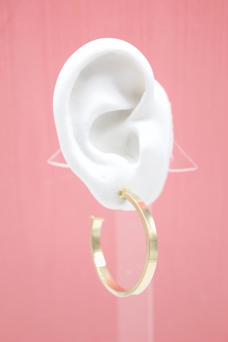 A white mannequin ear displays a stylish flat gold hoop earring against a pink background.