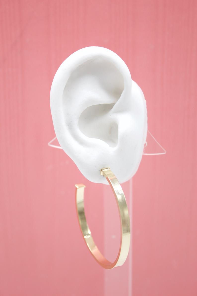 A white mannequin ear displays a large, flat gold hoop earring against a pink background.