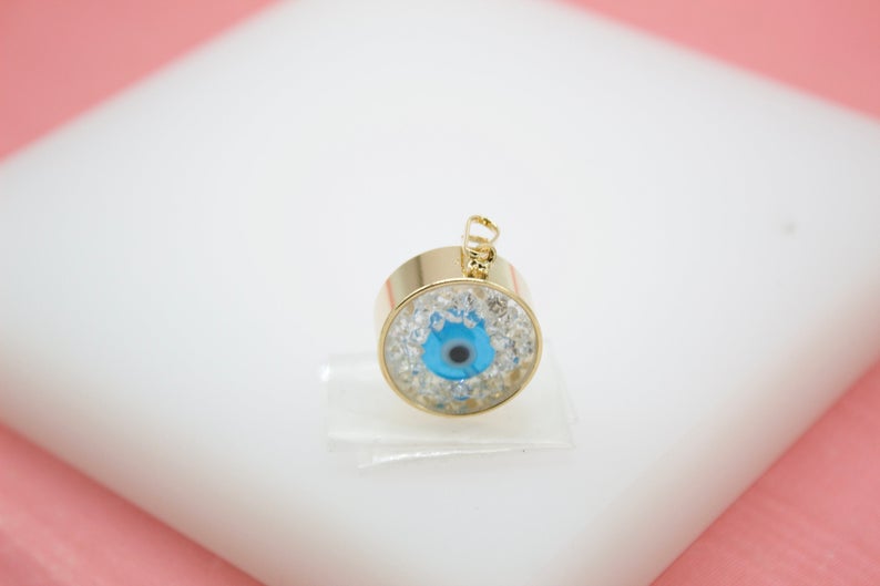 A gold evil eye pendant rests on a white display stand against a soft pink background.