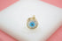 A gold evil eye pendant rests on a white display stand against a soft pink background.