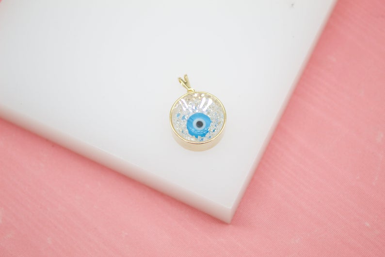 A gold-framed evil eye pendant rests on a white block against a pink background.