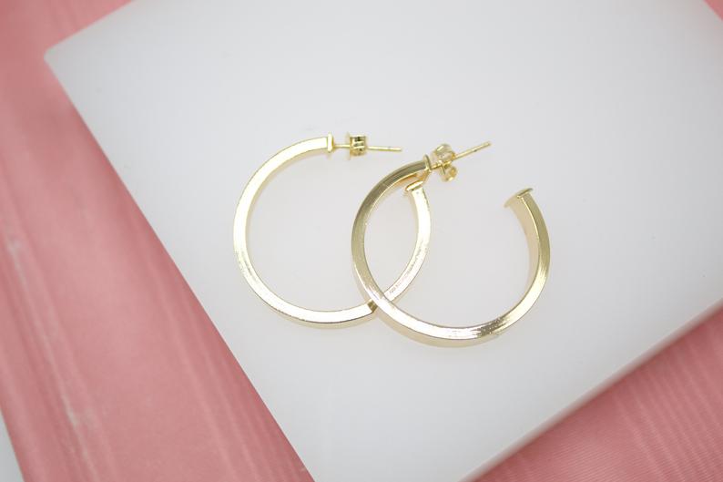 A pair of elegant gold hoop earrings rests gracefully on a white display surface against a soft pink background.