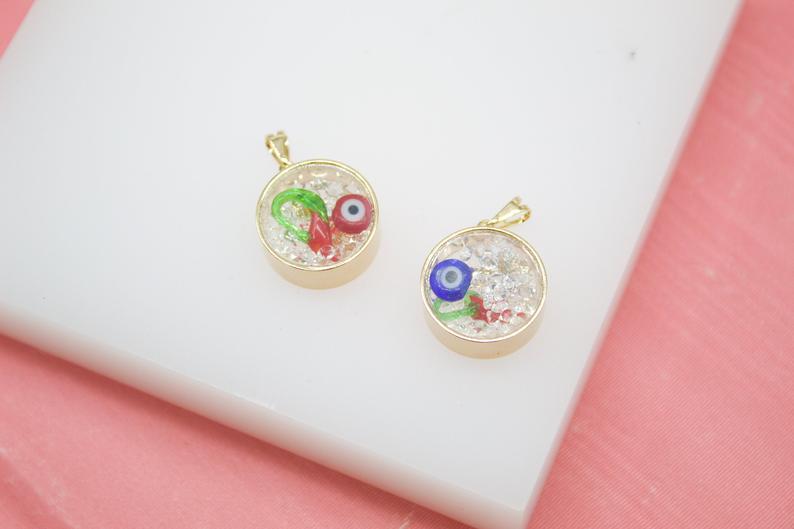Two gold-rimmed resin pendants filled with tiny white crystals and miniature evil eye charms rest on a white display surface.