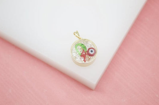 A gold-rimmed pendant rests on a white surface, showcasing a tiny red chili pepper and a protective blue evil eye charm set in clear beads.
