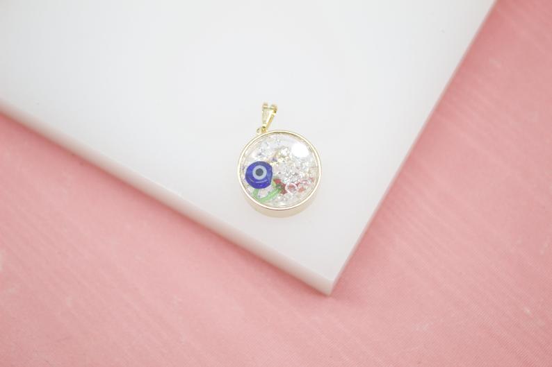 A round gold-framed pendant filled with clear resin, shimmering glitter, and a tiny blue evil eye charm rests on a white surface.