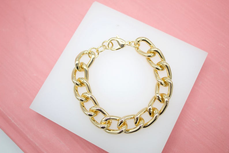 A thick gold chain link bracelet rests neatly on a white square surface against a soft pink background.
