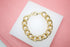 A thick gold chain link bracelet rests neatly on a white square surface against a soft pink background.