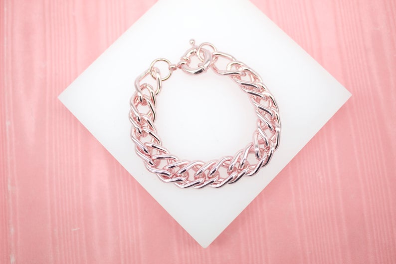 A thick rose gold chunky link bracelet rests on a white display pad against a pink textured background.