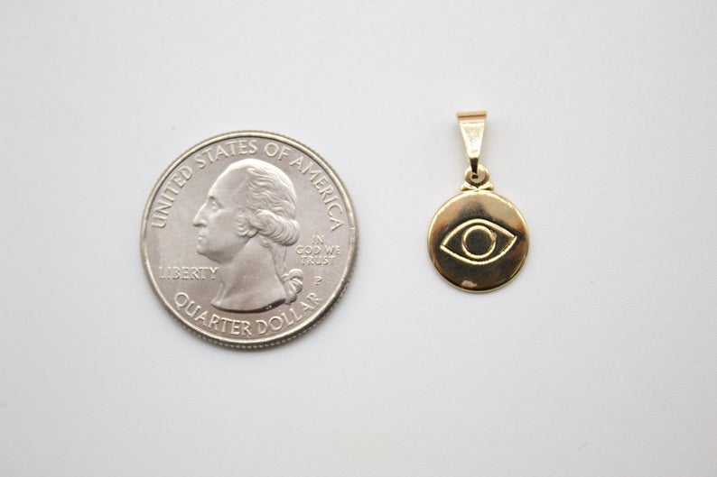 A small gold evil eye pendant rests next to a US quarter coin for size comparison on a white surface.