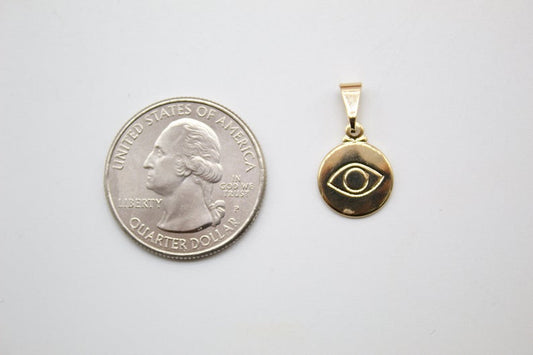 A small gold evil eye pendant rests next to a US quarter coin for size comparison on a white surface.