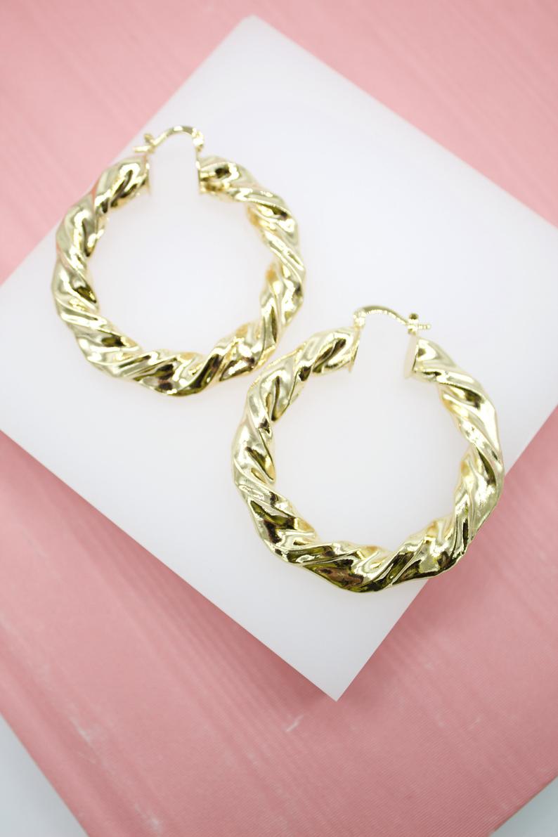 A pair of glossy gold twisted hoop earrings rests on a white display stand.