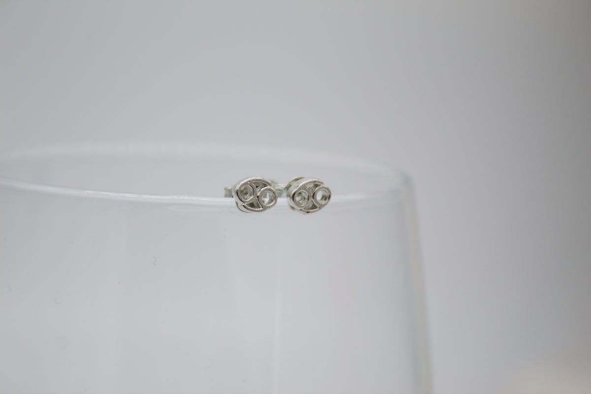 A pair of sterling silver Cancer zodiac sign stud earrings rests delicately on the rim of a clear glass.