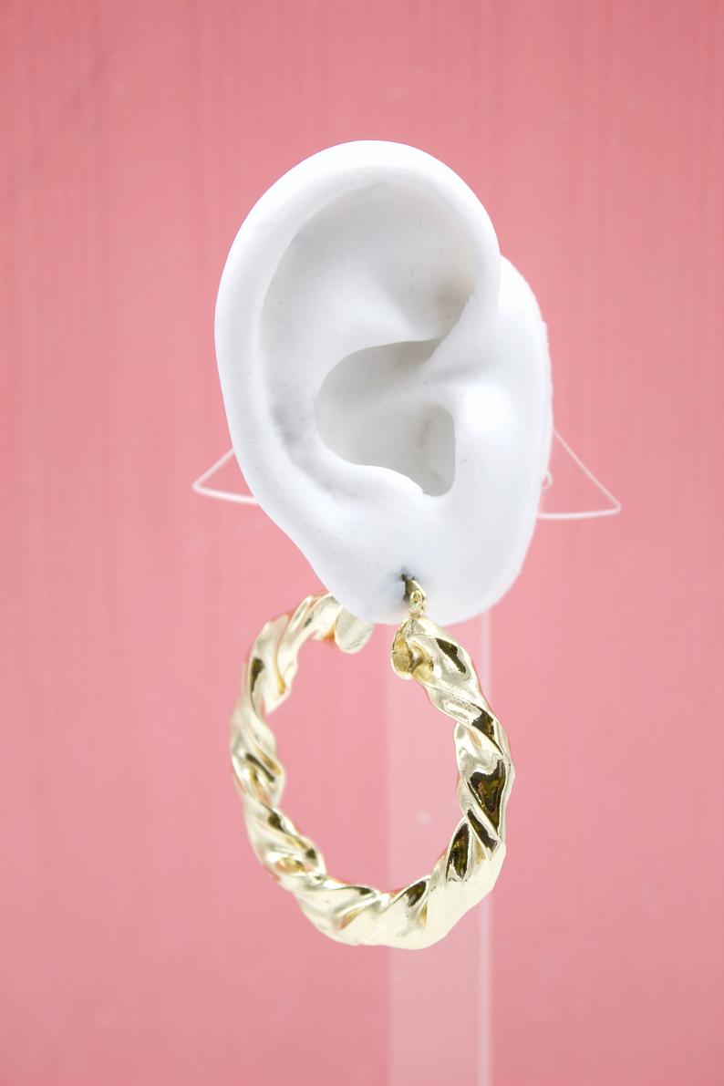 A white mannequin ear displays a shiny, twisted gold hoop earring against a soft pink background.