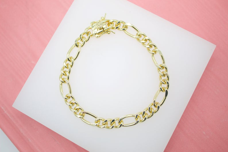 A gold Figaro chain link bracelet rests in a circle on a white display surface against a pink background.