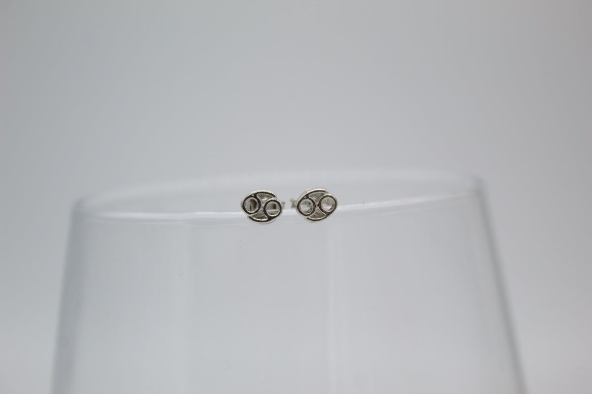 A pair of intricate silver stud earrings featuring the Cancer zodiac sign rest on the rim of a clear glass.