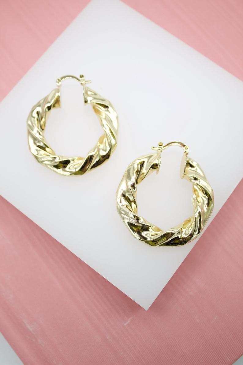 A pair of shiny gold twisted hoop earrings rests on a white square display against a pink background.