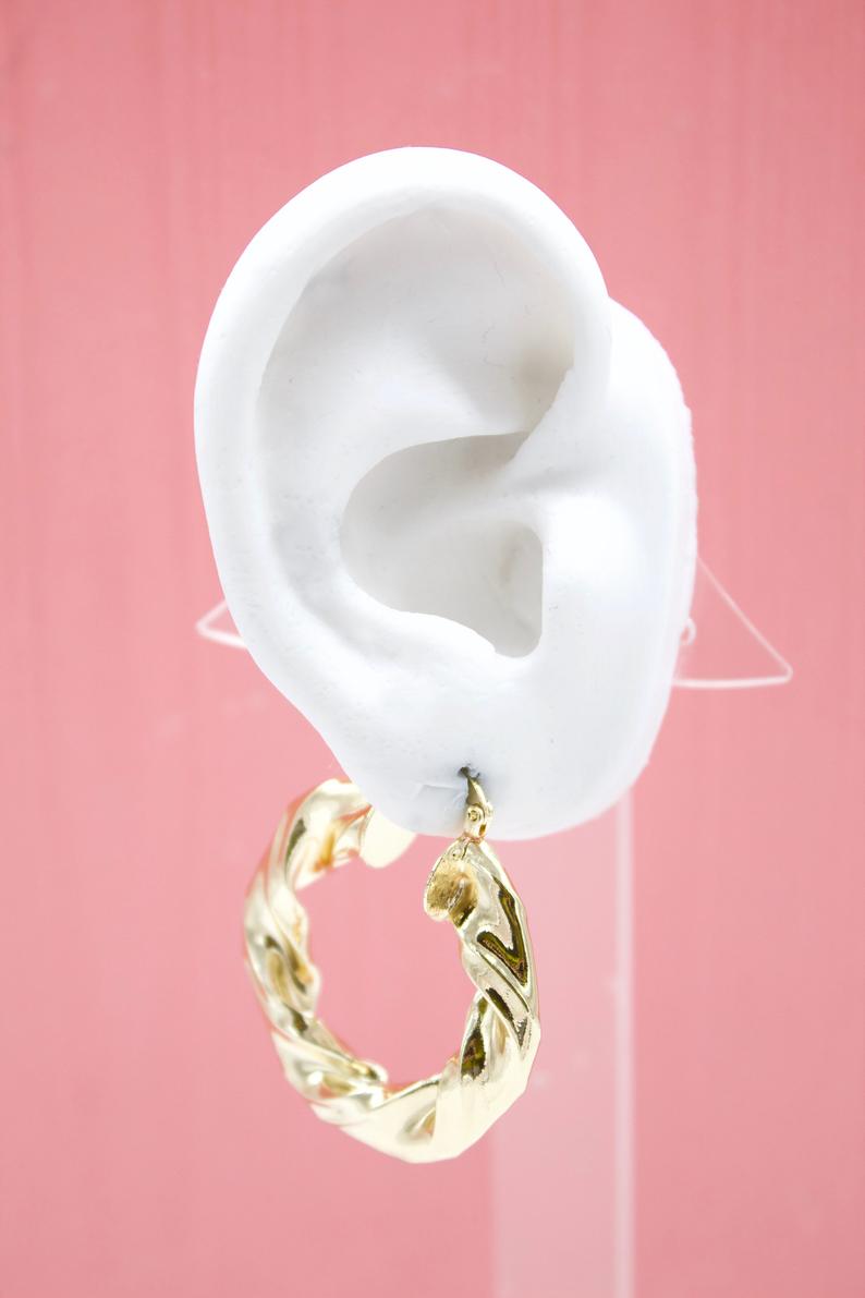 A white mannequin ear displays a chunky, twisted gold hoop earring against a soft pink background.
