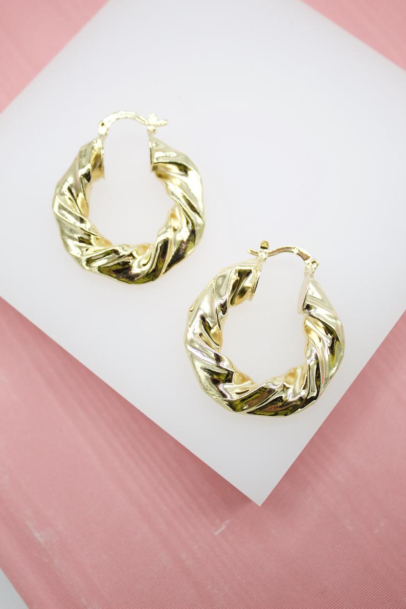 A pair of gleaming gold twisted hoop earrings rests on a white display surface against a pink background.