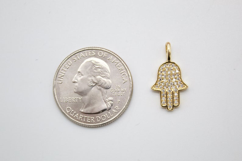A gold pavé diamond Hamsa hand pendant rests next to a US quarter coin to show its scale.