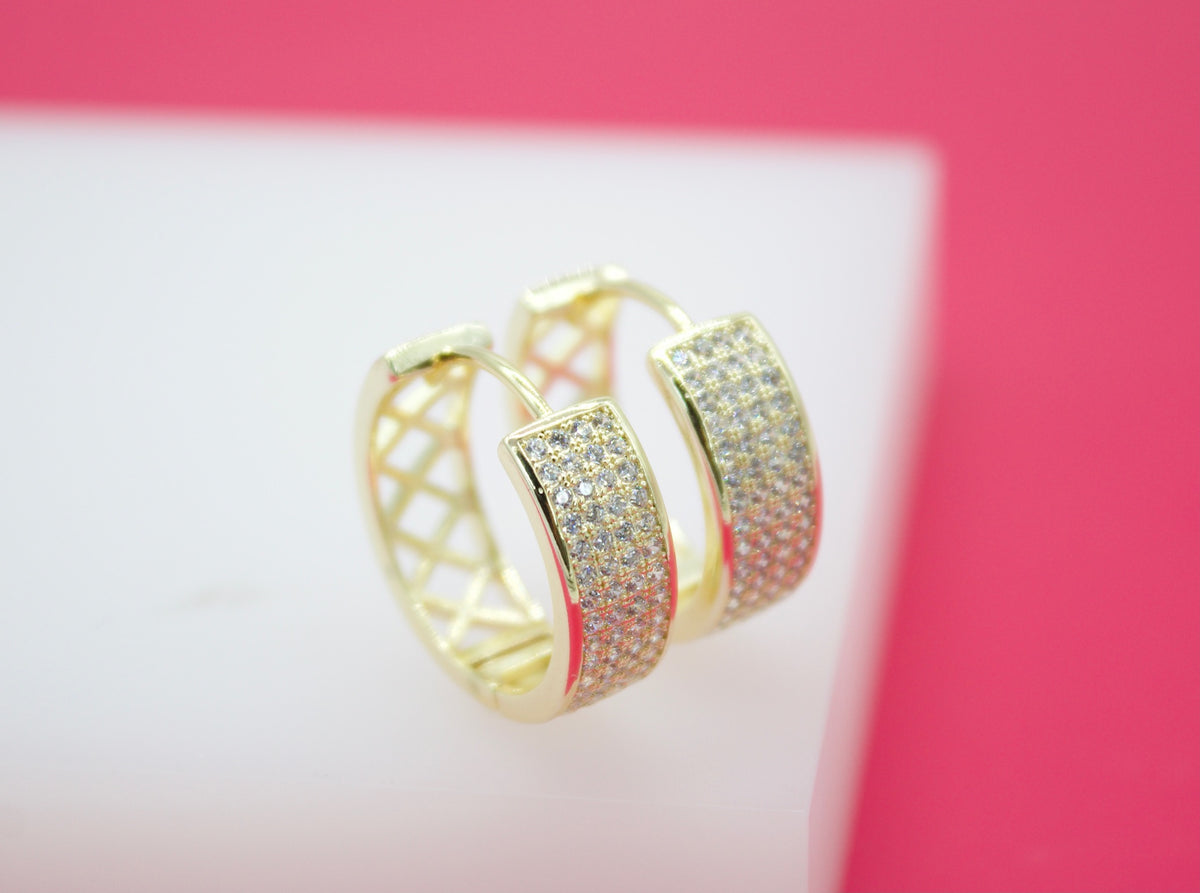 A pair of gold huggie hoop earrings encrusted with sparkling cubic zirconia pavé stones rests on a white display against a pink background.