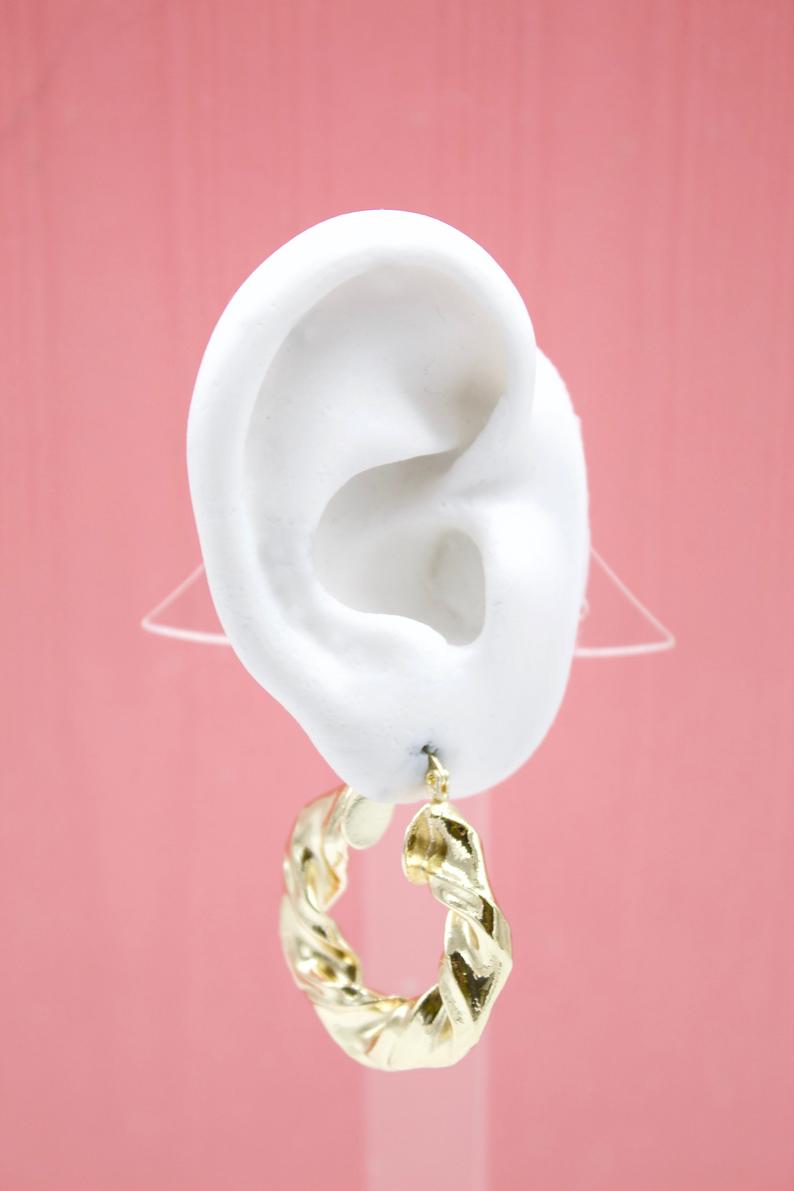 A white mannequin ear displays a chunky gold twisted hoop earring against a solid pink backdrop.