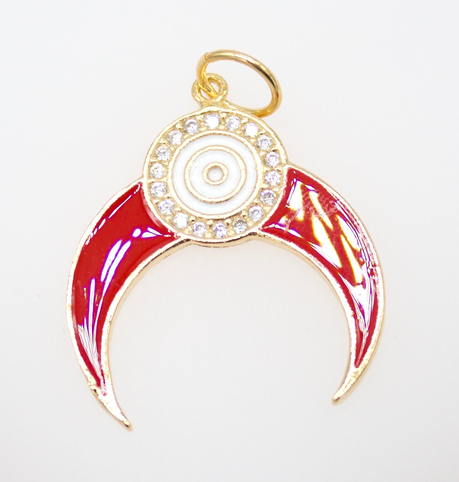 A gold-plated charm features a vibrant red enamel crescent shape accented by a sparkling rhinestone halo and a central white evil eye symbol.