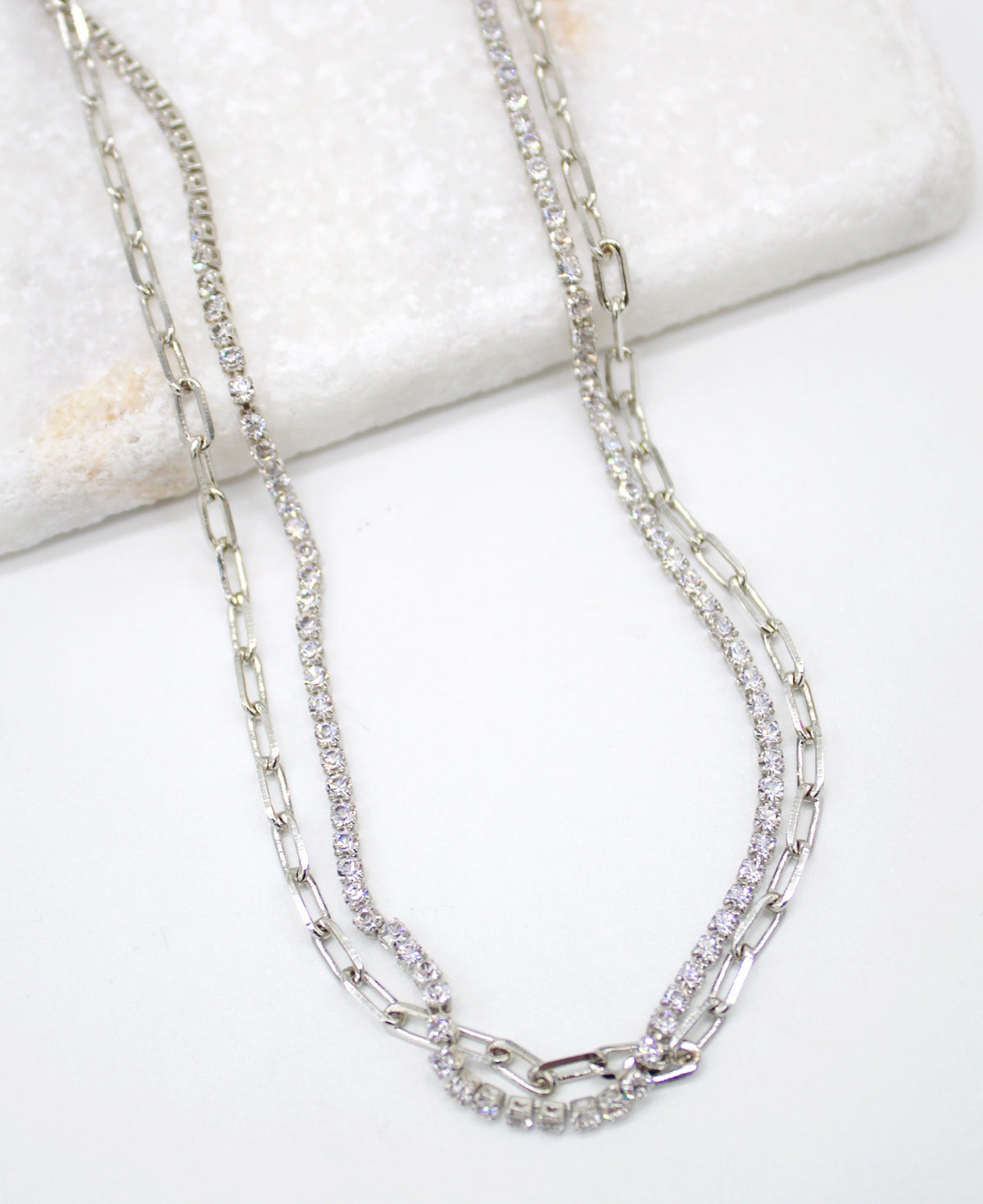 A layered silver chain necklace featuring a sparkling diamond tennis chain and a delicate link chain rests on a white surface.
