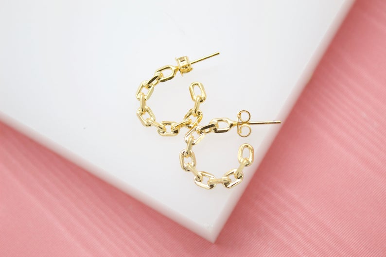 A pair of shiny gold chain link hoop earrings rests on a white display surface against a pink background.