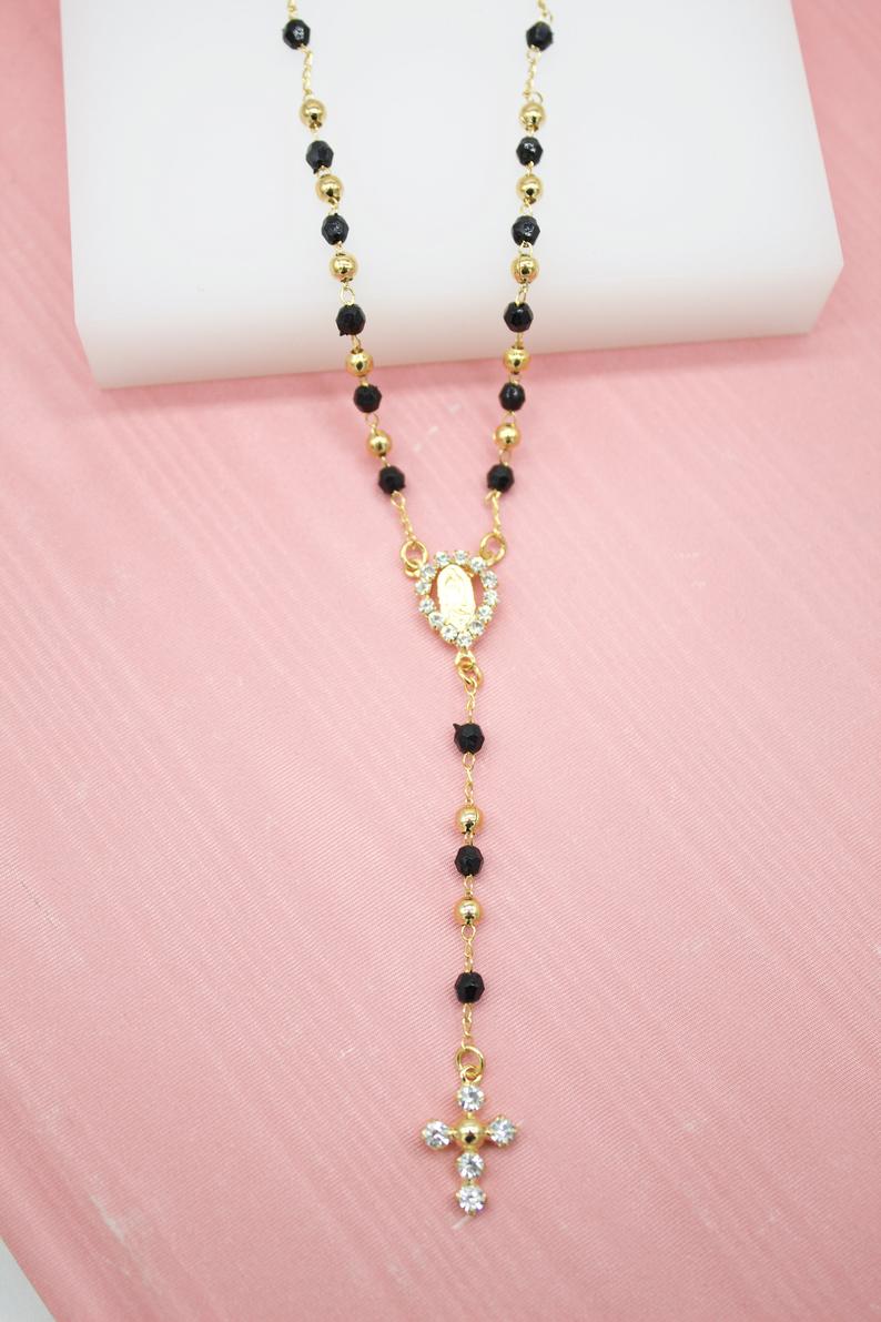 A gold-plated Catholic rosary necklace featuring black beads and a sparkling rhinestone crucifix rests against a soft pink backdrop.