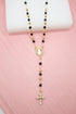 A gold-plated Catholic rosary necklace featuring black beads and a sparkling rhinestone crucifix rests against a soft pink backdrop.