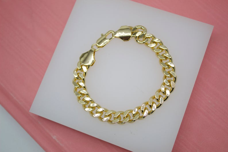 A polished gold curb chain bracelet rests on a white display pad against a textured pink background.
