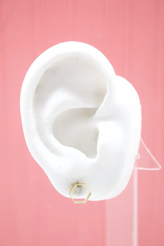 A white model ear displays a modern geometric gold hexagon stud earring against a soft pink background.