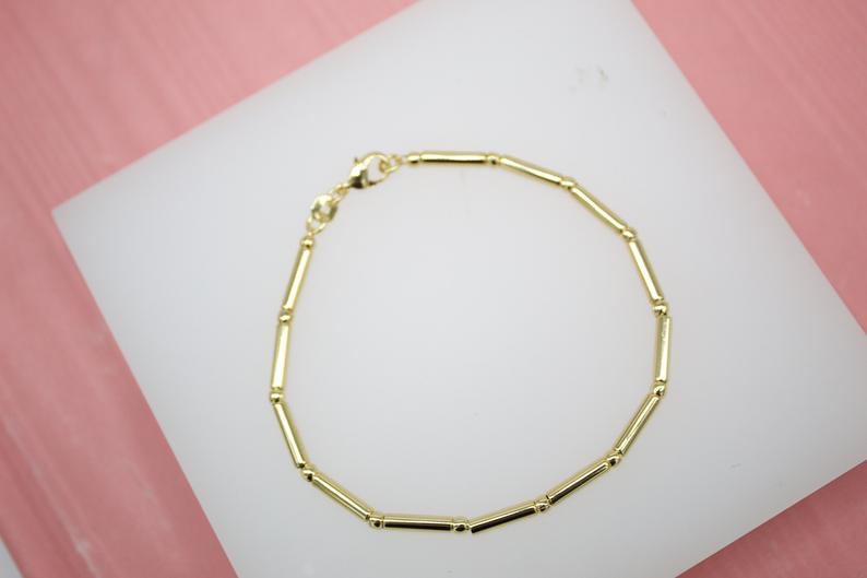 A delicate gold bar link bracelet rests on a white display surface against a pink background.