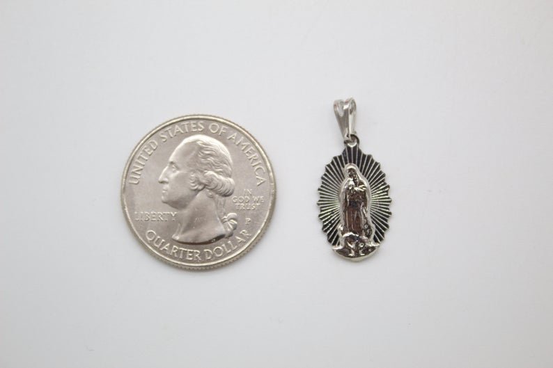 A silver Our Lady of Guadalupe pendant rests next to a US quarter coin for scale on a light background.